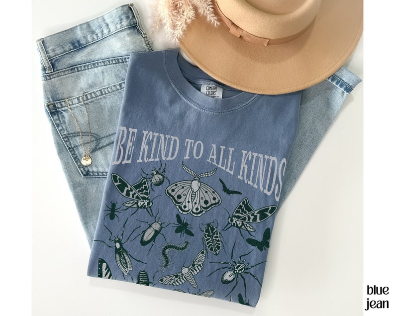 Op de afbeelding: Een blauw T-shirt met de tekst "BE KIND TO ALL KINDS" en insectenillustraties. Het shirt wordt gecombineerd met een spijkerbroek en een beige hoed. De afbeelding heeft een casual, ontspannen uitstraling.