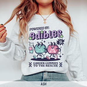 May include: A gray sweatshirt with a graphic of two cartoon gummies. The gummies are green and pink and have the word "THC" on them. The text on the sweatshirt reads "Powered by: edibles Save your lungs Garden gummies to the rescue".