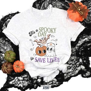 Puede incluir: Camiseta gris claro con el texto "It's a SPOOKY DAY to SAVE LIVES" y gr&aacute;ficos tem&aacute;ticos de Halloween. El dise&ntilde;o incluye un murci&eacute;lago, una calabaza y un fantasma con gorro de enfermera. Calabazas decorativas y tela negra rodean la camiseta.