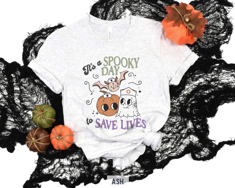 Puede incluir: Camiseta gris claro con el texto "It's a SPOOKY DAY to SAVE LIVES" y gr&aacute;ficos tem&aacute;ticos de Halloween. El dise&ntilde;o incluye un murci&eacute;lago, una calabaza y un fantasma con gorro de enfermera. Calabazas decorativas y tela negra rodean la camiseta.
