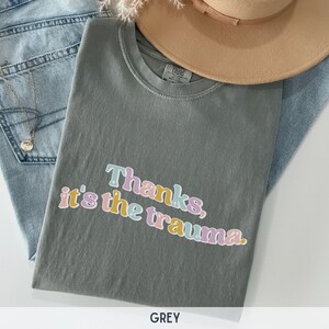 May include: A grey t-shirt with the text "Thanks, it's the trauma." printed in colorful letters.
