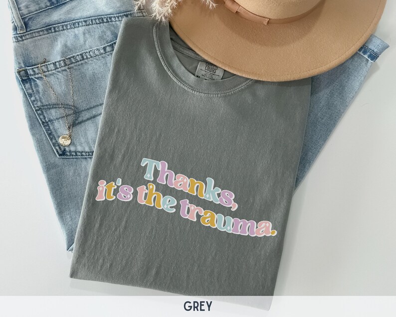 May include: A grey t-shirt with the text "Thanks, it's the trauma." printed in colorful letters.