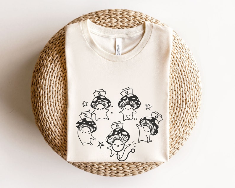 Op de afbeelding: Een wit T-shirt met een zwart-witte afbeelding van paddenstoelen die verpleegstersmutsen dragen en stethoscopen vasthouden. De tekst "Shroom Queens" is op het shirt gedrukt.