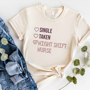 May include: A beige t-shirt with a graphic that says "Single, Taken, Night Shift Nurse" with a checkmark next to the last option.