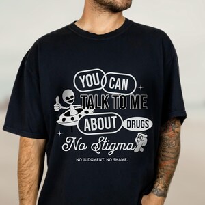 Peut inclure: T-shirt noir avec un motif graphique blanc repr&eacute;sentant un extraterrestre dans une soucoupe volante. Le texte dit "You Can Talk To Me About Drugs No Stigma No Judgement. No Shame."