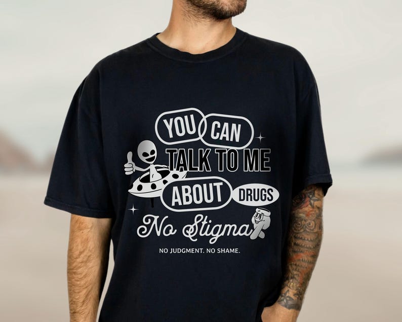 Peut inclure: T-shirt noir avec un motif graphique blanc repr&eacute;sentant un extraterrestre dans une soucoupe volante. Le texte dit "You Can Talk To Me About Drugs No Stigma No Judgement. No Shame."