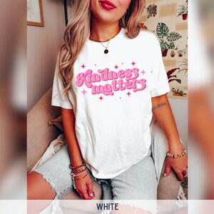 Pode incluir: T-shirt branco com a frase "Kindness matters" em rosa, com um estilo retro e estrelas. Uma pe&ccedil;a casual para uso di&aacute;rio.