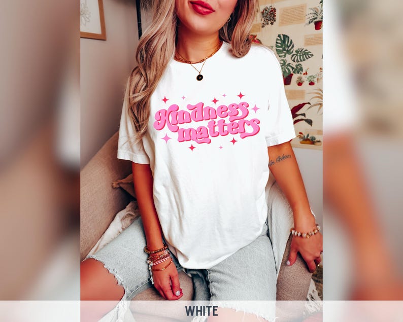 Pode incluir: T-shirt branco com a frase "Kindness matters" em rosa, com um estilo retro e estrelas. Uma pe&ccedil;a casual para uso di&aacute;rio.