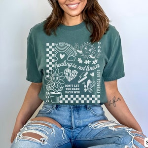 May include: A teal green t-shirt with a white graphic print featuring a checkered background, a flower, a rainbow, a sun, a heart, a lightning bolt, a butterfly, and the text "BE YOUR OWN SUNSHINE", "healing is not linear", "FIND JOY IN THE JOURNEY", "DON'T LET THE HARD DAYS WIN", "LOVE YOURSELF", and "988".