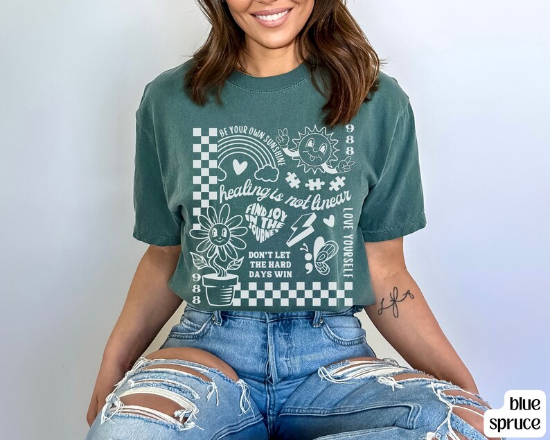May include: A teal green t-shirt with a white graphic print featuring a checkered background, a flower, a rainbow, a sun, a heart, a lightning bolt, a butterfly, and the text "BE YOUR OWN SUNSHINE", "healing is not linear", "FIND JOY IN THE JOURNEY", "DON'T LET THE HARD DAYS WIN", "LOVE YOURSELF", and "988".