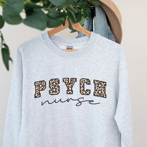 May include: Ash-colored sweatshirt with the words "PSYCH nurse" in a combination of leopard print and script lettering. The sweatshirt is hanging on a wooden hanger.