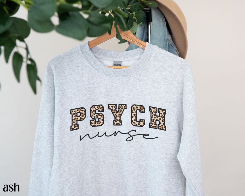 May include: Ash-colored sweatshirt with the words "PSYCH nurse" in a combination of leopard print and script lettering. The sweatshirt is hanging on a wooden hanger.