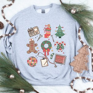 May include: A gray sweatshirt with a Christmas-themed design featuring medical symbols. The design includes a gingerbread person, a stethoscope, a candy cane, a Christmas tree, a wreath, a stocking, and a heart monitor.