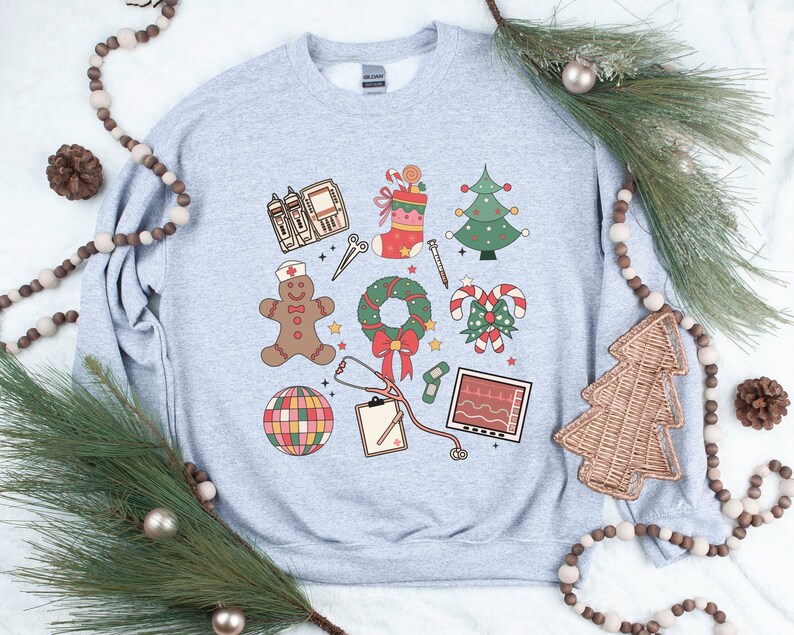 May include: A gray sweatshirt with a Christmas-themed design featuring medical symbols. The design includes a gingerbread person, a stethoscope, a candy cane, a Christmas tree, a wreath, a stocking, and a heart monitor.