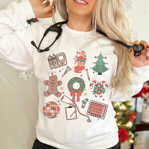 May include: White sweatshirt with a festive Christmas design featuring a gingerbread person, a wreath, candy canes, a stocking, a Christmas tree, a stethoscope, a medical monitor, and a medical IV bag.