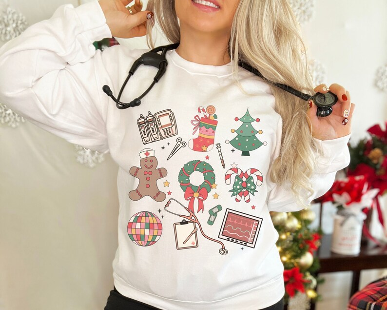 May include: White sweatshirt with a festive Christmas design featuring a gingerbread person, a wreath, candy canes, a stocking, a Christmas tree, a stethoscope, a medical monitor, and a medical IV bag.