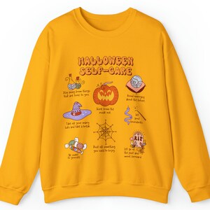 May include: Yellow sweatshirt with a Halloween self-care graphic. The graphic features a pumpkin, a witch's hat, a spiderweb, candy, and other Halloween-themed illustrations. The text on the graphic reads "Halloween Self-Care".