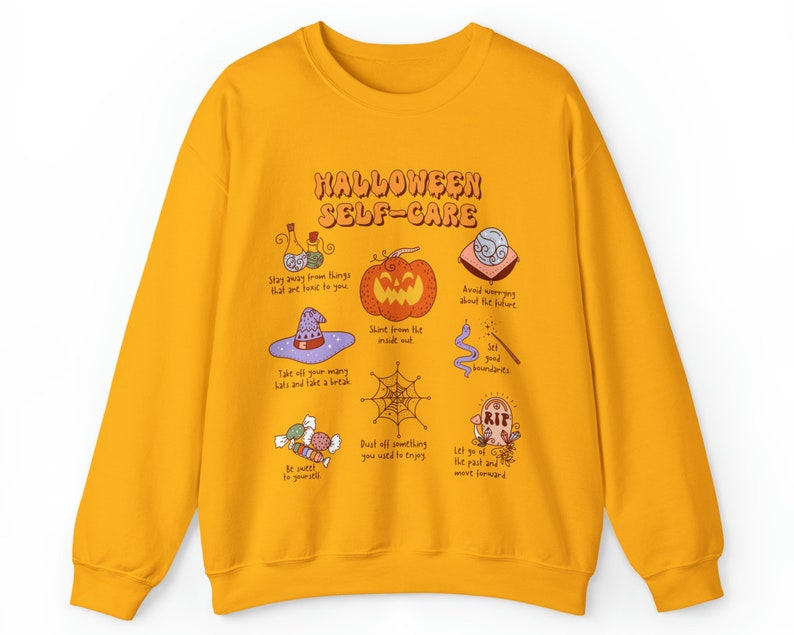 May include: Yellow sweatshirt with a Halloween self-care graphic. The graphic features a pumpkin, a witch's hat, a spiderweb, candy, and other Halloween-themed illustrations. The text on the graphic reads "Halloween Self-Care".