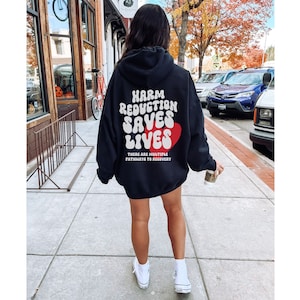Peut inclure: Un sweat &agrave; capuche noir avec le texte "HARM REDUCTION SAVES LIVES" en blanc, avec un c&oelig;ur rouge. Le sweat &agrave; capuche porte &eacute;galement le texte "THERE ARE MULTIPLE PATHWAYS TO RECOVERY". La personne porte des baskets blanches.