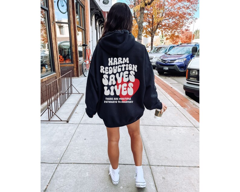 Peut inclure: Un sweat &agrave; capuche noir avec le texte "HARM REDUCTION SAVES LIVES" en blanc, avec un c&oelig;ur rouge. Le sweat &agrave; capuche porte &eacute;galement le texte "THERE ARE MULTIPLE PATHWAYS TO RECOVERY". La personne porte des baskets blanches.