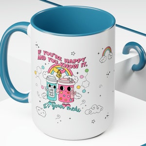 May include: White ceramic mug with a teal blue handle and rim. The mug features a cartoon illustration of two pill bottles with faces and the text "If you're happy and you know it, it's your meds".
