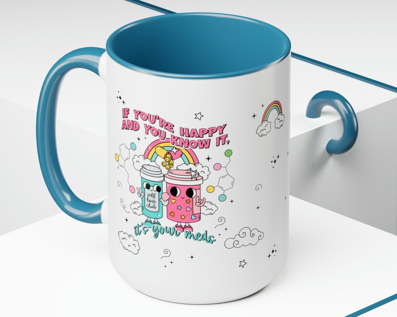 May include: White ceramic mug with a teal blue handle and rim. The mug features a cartoon illustration of two pill bottles with faces and the text "If you're happy and you know it, it's your meds".