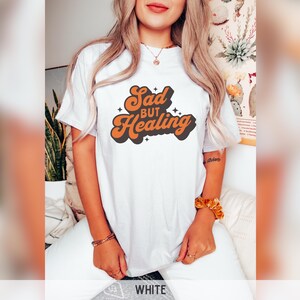 Puede incluir: Camiseta blanca con el texto "Sad but Healing" en fuente retro naranja y negro. La camiseta es de estilo casual, adecuada para el uso diario. El dise&ntilde;o presenta una est&eacute;tica vintage.