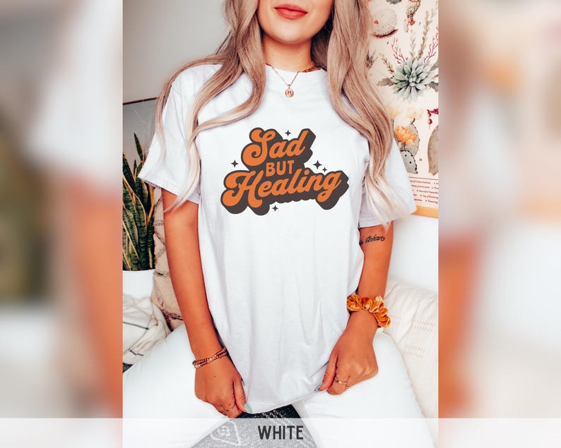 Puede incluir: Camiseta blanca con el texto "Sad but Healing" en fuente retro naranja y negro. La camiseta es de estilo casual, adecuada para el uso diario. El dise&ntilde;o presenta una est&eacute;tica vintage.
