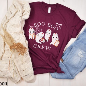 May include: A maroon t-shirt featuring a white graphic of three ghosts dressed as nurses. The ghosts are holding medical equipment and the text "Boo Boo Crew" is above them.