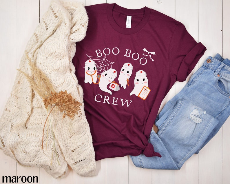 May include: A maroon t-shirt featuring a white graphic of three ghosts dressed as nurses. The ghosts are holding medical equipment and the text "Boo Boo Crew" is above them.