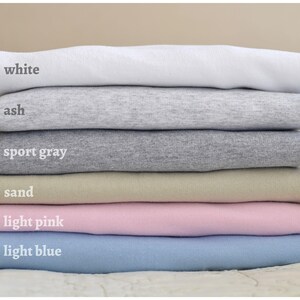 May include: Stack of six t-shirts in different colors: white, ash, sport gray, sand, light pink, and light blue.