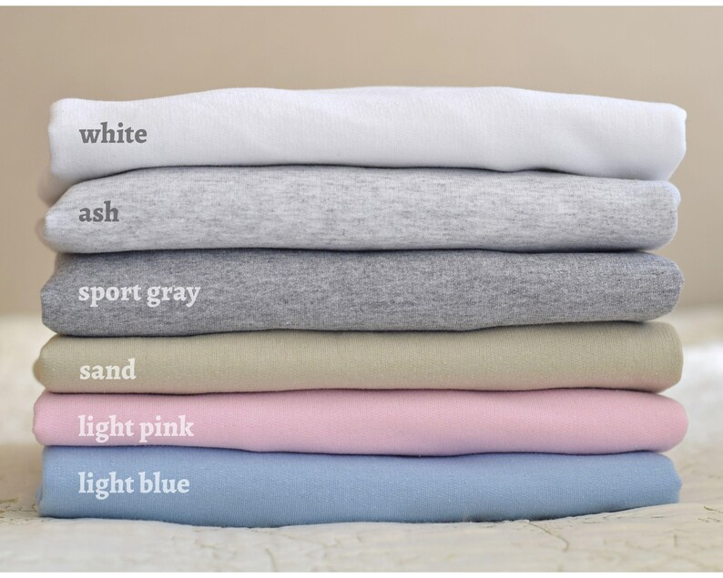 May include: Stack of six t-shirts in different colors: white, ash, sport gray, sand, light pink, and light blue.