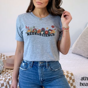 May include: A gray t-shirt with a floral design and the text "NEUROSCIENCE" in black. The model is wearing blue jeans.
