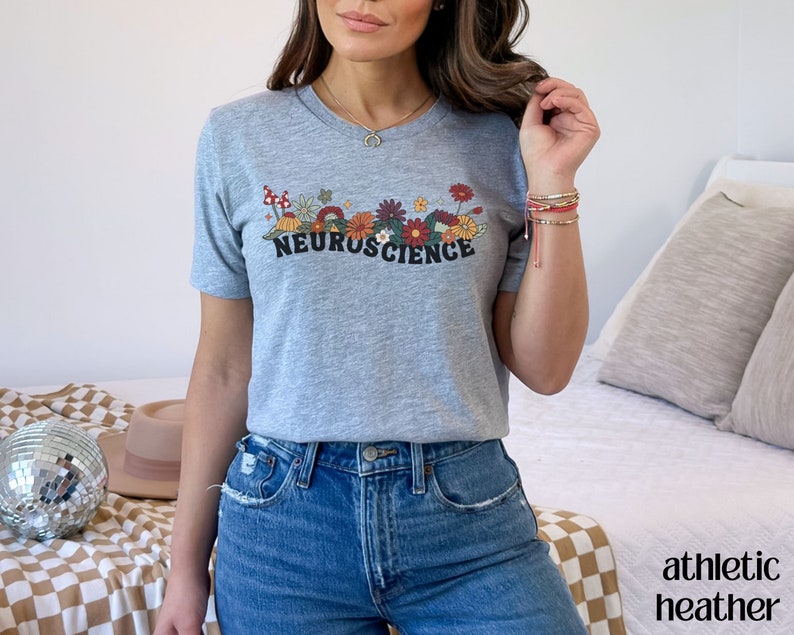 May include: A gray t-shirt with a floral design and the text "NEUROSCIENCE" in black. The model is wearing blue jeans.