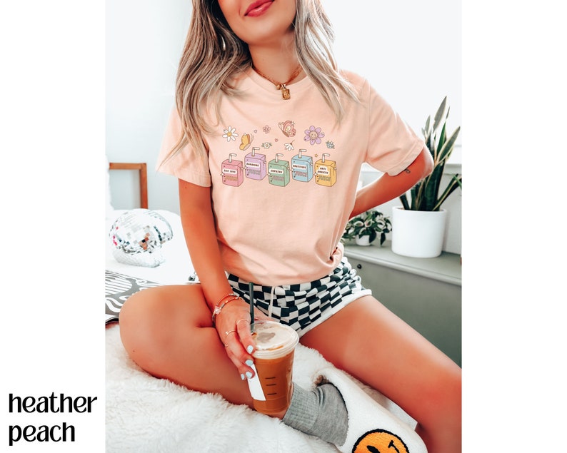 May include: A pink t-shirt with a graphic of colorful juice boxes and flowers. The shirt is worn by a person sitting on a white fuzzy surface, holding a cup of coffee.