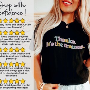 May include: Black t-shirt with the text "Thanks, it's the trauma." in colorful letters. The shirt is being worn by a person with long blonde hair. The shirt has received five-star reviews from customers who praise the quality and design.