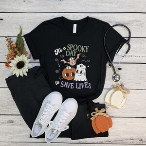 Puede incluir: Camiseta negra con el texto "It's a Spooky Day to Save Lives" y un gr&aacute;fico tem&aacute;tico de Halloween. El gr&aacute;fico incluye un murci&eacute;lago, una calabaza y un fantasma. La camiseta se combina con pantalones negros y zapatillas blancas. Tambi&eacute;n hay un estetoscopio y calabazas decorativas.