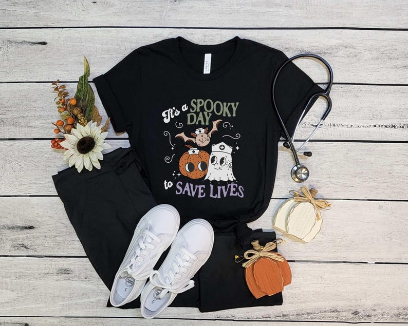 Puede incluir: Camiseta negra con el texto "It's a Spooky Day to Save Lives" y un gr&aacute;fico tem&aacute;tico de Halloween. El gr&aacute;fico incluye un murci&eacute;lago, una calabaza y un fantasma. La camiseta se combina con pantalones negros y zapatillas blancas. Tambi&eacute;n hay un estetoscopio y calabazas decorativas.