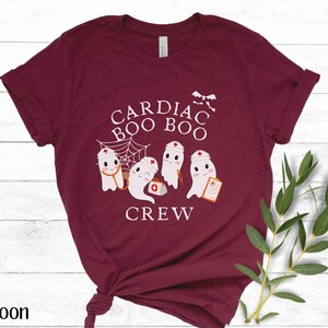 May include: A maroon t-shirt with a white graphic of four ghosts wearing nurse hats and holding medical equipment. The text "Cardiac Boo Boo Crew" is printed on the shirt.