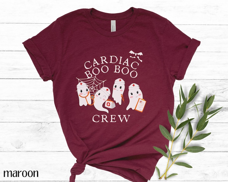 May include: A maroon t-shirt with a white graphic of four ghosts wearing nurse hats and holding medical equipment. The text "Cardiac Boo Boo Crew" is printed on the shirt.