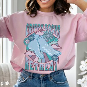 May include: Light pink sweatshirt with a graphic design of blue socks with a white pattern, flowers, and the text "Grippy Socks Retreat".