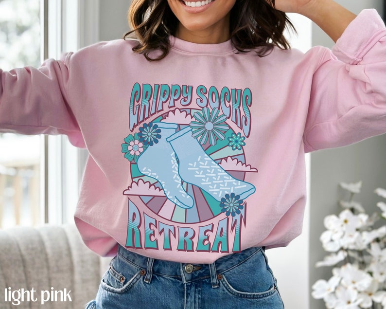 May include: Light pink sweatshirt with a graphic design of blue socks with a white pattern, flowers, and the text "Grippy Socks Retreat".