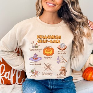 May include: A beige sweatshirt with a graphic print of a pumpkin, a witch's hat, a spiderweb, and other Halloween-themed illustrations. The text on the sweatshirt reads "Halloween Self-Care".