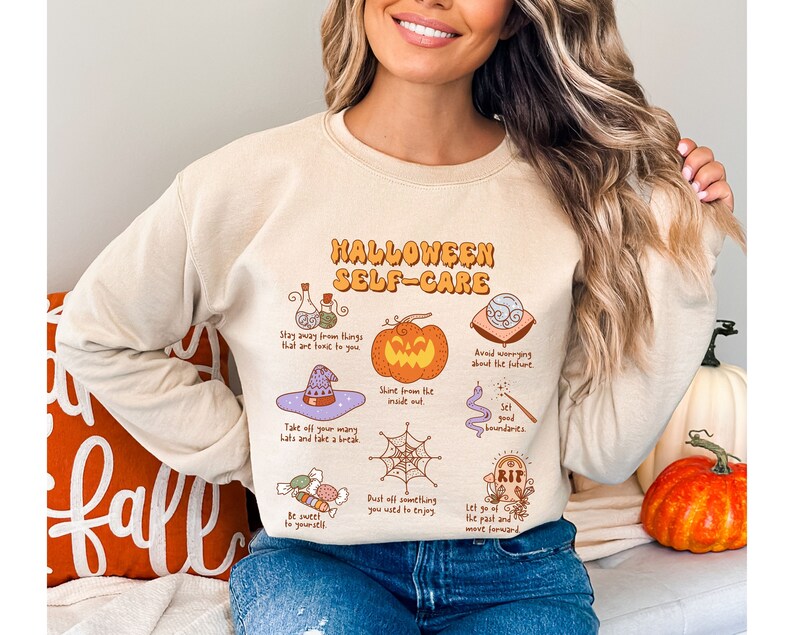 May include: A beige sweatshirt with a graphic print of a pumpkin, a witch's hat, a spiderweb, and other Halloween-themed illustrations. The text on the sweatshirt reads "Halloween Self-Care".