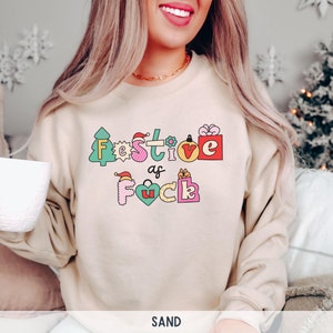 Op de afbeelding: Een beige sweatshirt met een feestelijk ontwerp dat "Festive as Fuck" zegt in kleurrijke, omrande letters. De letters zijn versierd met kerstthema-pictogrammen zoals een kerstboom, een cadeaudoos en een kerstmanmuts.