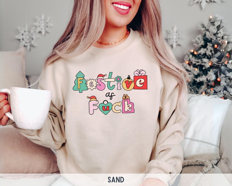 Op de afbeelding: Een beige sweatshirt met een feestelijk ontwerp dat "Festive as Fuck" zegt in kleurrijke, omrande letters. De letters zijn versierd met kerstthema-pictogrammen zoals een kerstboom, een cadeaudoos en een kerstmanmuts.