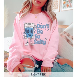 Op de afbeelding: Een lichtroze sweatshirt met een afbeelding van een cartoon IV-zak met de tekst "Don't Be So Salty".