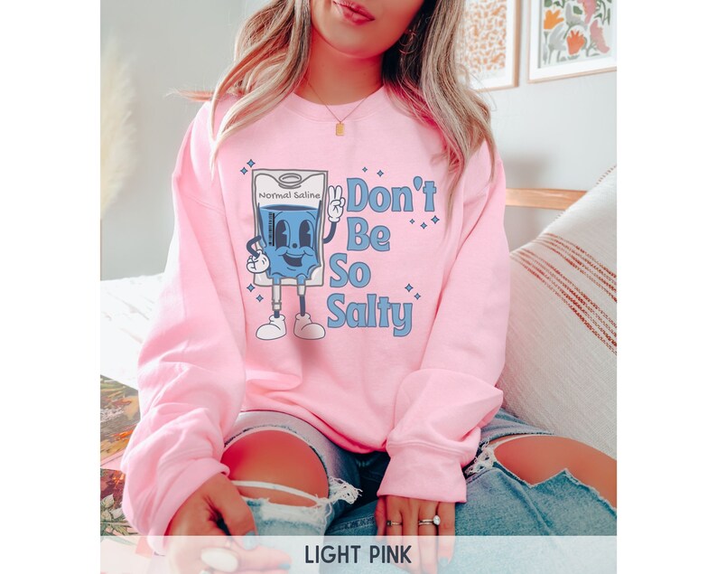 Op de afbeelding: Een lichtroze sweatshirt met een afbeelding van een cartoon IV-zak met de tekst "Don't Be So Salty".