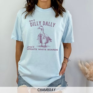 May include: Light blue t-shirt with a graphic of a duck wearing a doctor's coat and stethoscope. The text on the shirt reads "born to dilly dally forced to update white boards".