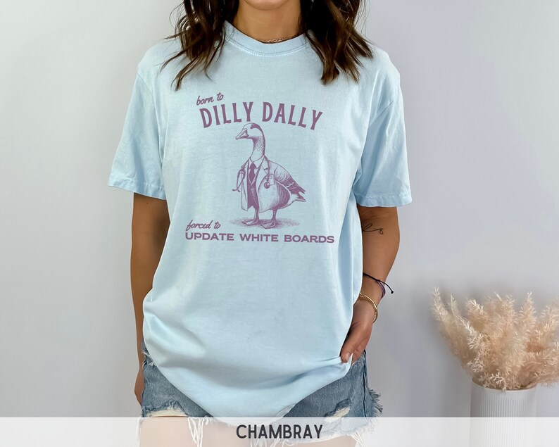 May include: Light blue t-shirt with a graphic of a duck wearing a doctor's coat and stethoscope. The text on the shirt reads "born to dilly dally forced to update white boards".